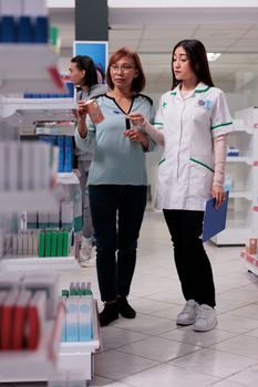 Customer and pharmacist examining medical products on shelves, giving advice in buying box of supplements or vitamins. Asian people talking about healthcare treatment and bottles of pills.