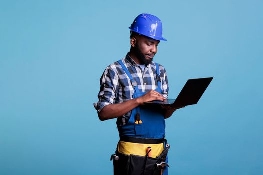 Construction worker in work uniform and hard hat using modern laptop computer. Professional male construction site supervisor wearing a tool belt while using a computer on a blue background.