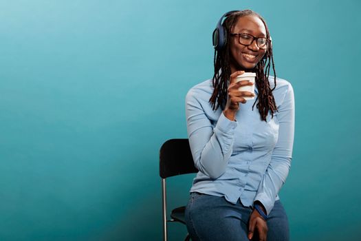 Cheerful happy african american woman smiling heartily while listening to music playlist on wireless headphones. Joyful positive young adult person with earphones enjoying radio on blue background.