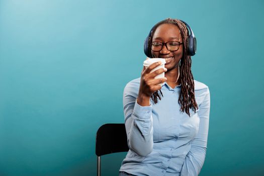 Joyful positive young adult person with earphones enjoying radio while sipping coffee. Cheerful happy african american woman smiling heartily while listening to music playlist on wireless headphones.