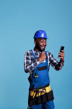 African american builder excited by news of new construction contract received by video call on cell phone. Contractor celebrating with emotion holding mobile device against blue background.