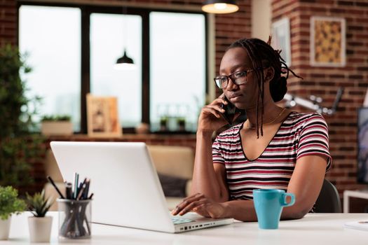 Freelancer talking on smartphone with client, working on laptop, in home office. Busy remote worker answering phone call, employee in glasses having conversation on telephone