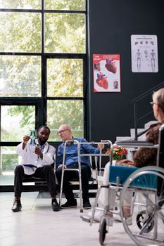 Injured senior patient sitting in hospital area analyzing lungs radiography with practitioner doctor discussing medical diagnosis during examination. Patients waiting in reception area