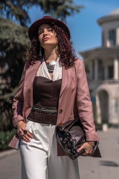 Woman park city. Stylish woman in a hat walks in a park in the city. Dressed in white corset trousers and a pink jacket with a bag in her hands