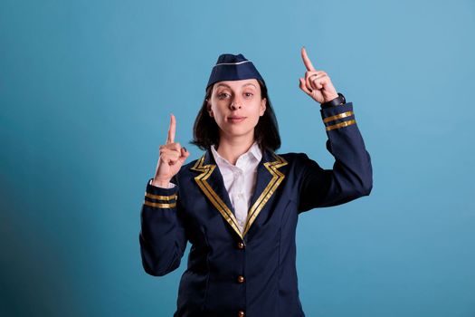Stewardess pointing up with index fingers, aviation academy flight attendant advertising product, medium shot. Confident air hostess showing upwards with forefinger in studio with blue background
