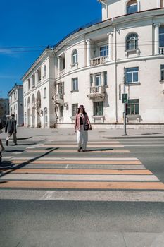 Woman city road crossing. Stylish woman in a hat crosses the road at a pedestrian crossing in the city. Dressed in white trousers and a jacket with a bag in her hands