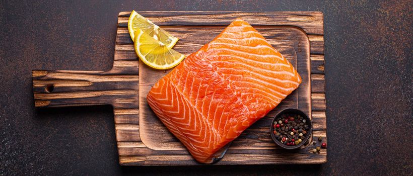 Fresh raw salmon fillet on wooden kitchen cutting board with seasonings and lemon top view on dark brown rustic background