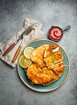 Crispy panko breaded fried chicken fillet with green salad and lemon cut on plate on gray rustic concrete background table with ketchup from above. Japanese style deep fried coated chicken breasts.