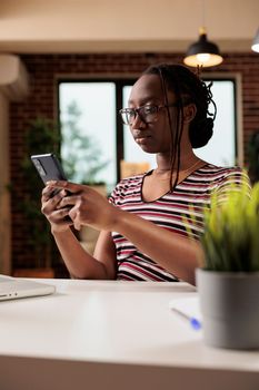 Young african american woman typing message on smartphone, chatting in social media at workplace. Freelancer surfing internet on mobile phone, sitting at desk in home office