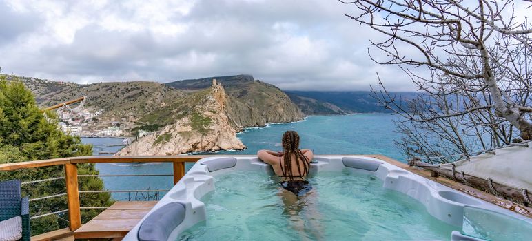 Outdoor jacuzzi with mountain and sea views. A woman in a black swimsuit is relaxing in the hotel pool, admiring the view.