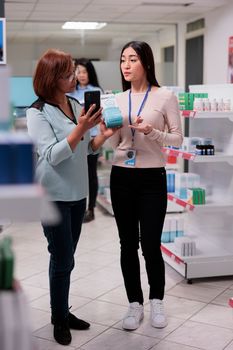 Pharmacist working in drugstore with elderly client, helping woman to find prescription drugs box on pharmacy shelves. Customer asking specialist about pills in pharmaceutical drugstore.