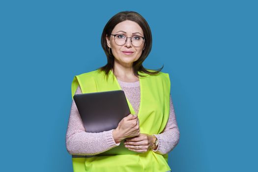 Portrait of engineer manager woman in helmet vest holding laptop, over blue studio background. Logistics, construction, industry, management, architecture, engineering, staff workers concept