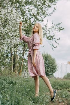 Blonde girl on a spring walk in the garden with cherry blossoms. Female portrait, close-up. A girl in a pink polka dot dress