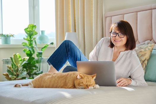 Middle aged woman at home on couch with laptop and cat. Mature 40s female looking at camera, on bed with pet. Work at home, technology, leisure, freelancing, lifestyle, people and animals concept