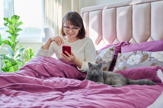 Middle-aged woman sitting in bed in morning looking at smartphone, with pet gray cat. Lifestyle, home, people animals concept