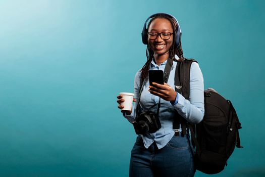 Joyful professional photographer with wireless headphones and modern smartphone listening to music on blue background. Happy woman with camera device enjoying songs while browsing internet on phone.
