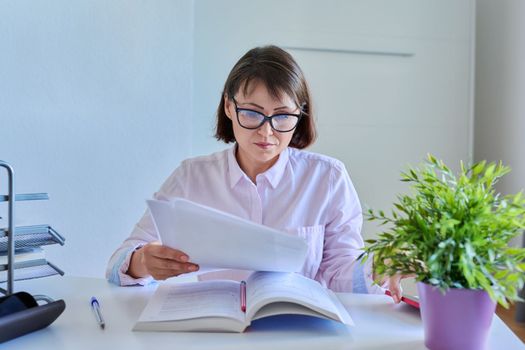 Middle-aged female working at home at a table with papers, books. Mature woman in glasses holding business papers documents, education, home workplace, paperwork concept