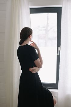 Silhouette of young woman standing by the window. Emotional portrait of a beautiful lonely girl with dark hair indoors.