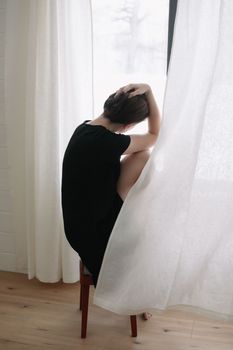 Silhouette of young woman sitting at the window, Dramatic emotional portrait of a beautiful lonely girl with dark hair indoors