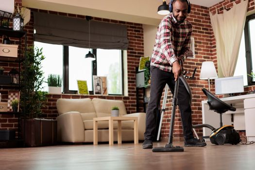 African american man vacuuming dust at home, spring cleaning on his day off from work. Using wireless headphones to listen to favorite music playlist while doing housework.