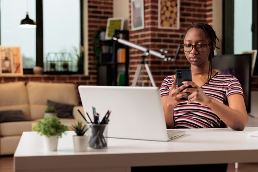 Young african american woman sending text messages on smartphone, communicating in social media network. Remote worker having break from work, surfing internet on mobile phone