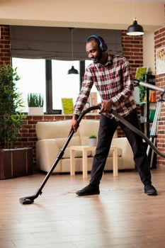 Smiling African american man with wireless headset cleaning floor at home with vacuum cleaner. Guy taking care of household chores, doing spring cleaning in urban apartment.