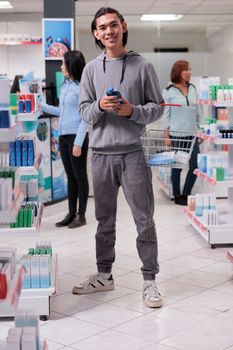 Asian male client coming at pharmacy shop to buy medication and prescription treatment, looking at boxes of health care products on shelves. Customer holding pharmaceutical supplies in drugstore.
