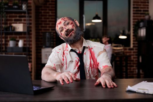 Portrait of dangerous zombie man sitting at desk, looking creepy and terrifiyng in company office. Brain eating cruel monster with bloody wounds working on laptop, horrible possessed devil.