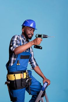 Construction worker on ladder drilling wall with cordless drill doing renovation of interior space. African american man in studio shot against blue background, construction and renovation concept.