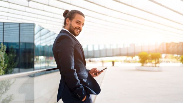 Businessman standing outdoor use smartphone looking camera screen and smile. Handsome hispanic male business person in business suit holding smartphone in hand. Modern business person low angle view