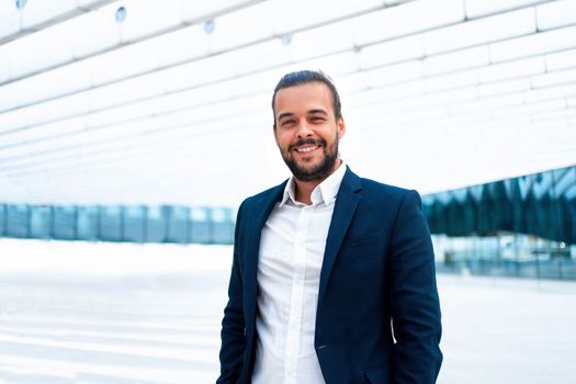 Successful businessman in suit with beard standing in front of office building looking at camera and smile. Hispanic male business person side view portrait. Free space. Modern business people concept