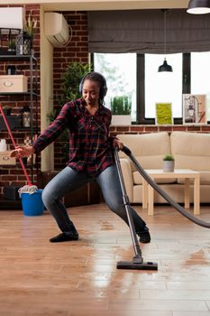 Positive housewife enjoying spring cleaning at home, singing while vacuuming in living room. Listening to music with wireless headphones relaxed looking cheerful, taking care of finishing household chores.