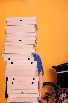 Pizzeria worker with blue uniform holding large stack of pizza delivering to client on bike. Food delivery employee standing in studio with yellow background. Food service and transportation