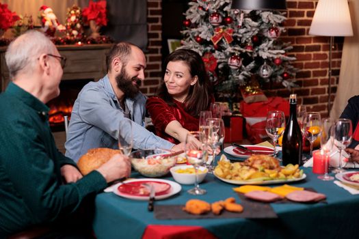 Romantic couple celebrating christmas with family, happy boyfriend and girlfriend talking at festive dinner table. Xmas celebration, wife and husband eating traditional meal with parents