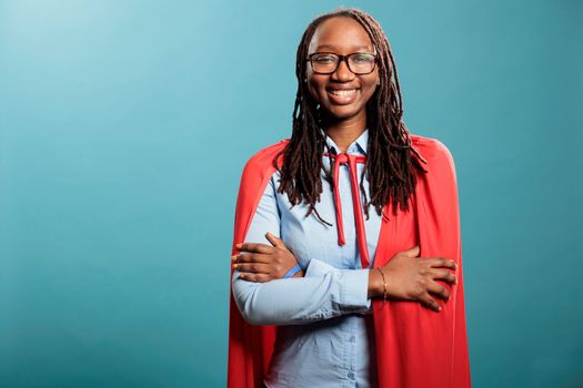 Joyful smiling heartily brave and selfless justice defender standing with arms crossed on blue background. Positive and happy young adult superhero woman wearing mighty hero red cloak. Studio shot