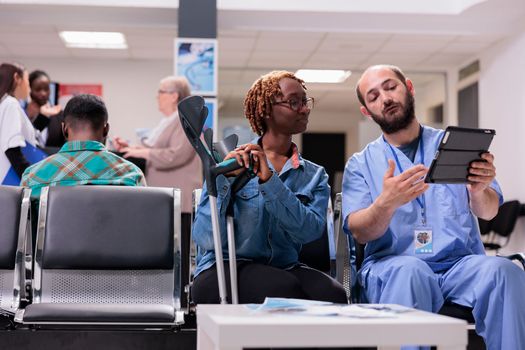 General practitioner interpreting clinical analysis of female young african american patient at sanatorium. Male nurse scheduling appointment for woman with walking difficulties in hospital lobby.