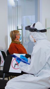 Stomatologist in protective suit reviewing registration form with patient explaining treatment writing on clipboard in covid-19 pandemic. Medical nurse wearing face shield, coverall, mask and gloves.