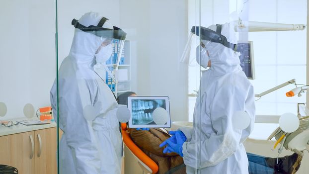 Slow motion close up of stomatological doctors with ppe suit analysing teeth x-ray using tablet in dental room, planning surgery during global pandemic while patient waiting on stomatological chair