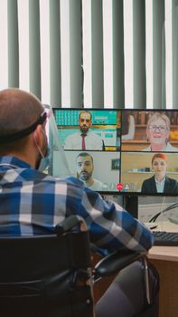 Paralysed manager in wheelchair talking with mask and visor during videocall having online conference in business office. Immobilized freelancer working in financial company respecting social distance