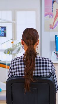 Assistant with face mask against covid-19 coming in reception typing on computer while patients sitting on chairs in waiting room talking keeping distance. Concept of new normal dentist visit.