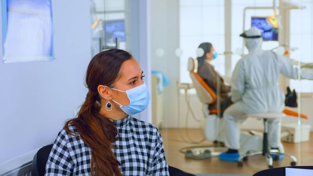 Portrait of young woman with protection mask discussing sitting on chairs keeping social distance in stomatological clinic, waiting for doctor during coronavirus. Concept of new normal dentist visit