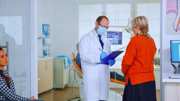 Orthodontist with mask speaking with elderly woman standing in waiting area of stomatological clinic taking notes on clipboard. Nurse typing on computer appointments in modern crowded office