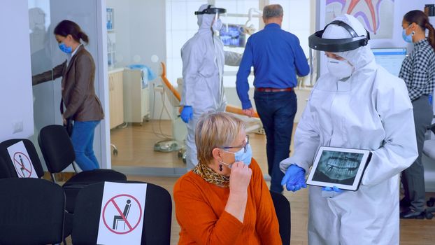 Dentist doctor with face shield pointing on tablet display explaining dental x-ray to senior patient during global pandemic. Nurse wearing protection suit, overall, mask and gloves, new normal