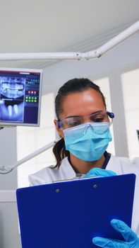 Dentistry doctor interrogating patient and taking notes on clipboard before surgery. Doctor and nurse working in modern orthodontic office, writing and examining person wearing protection mask