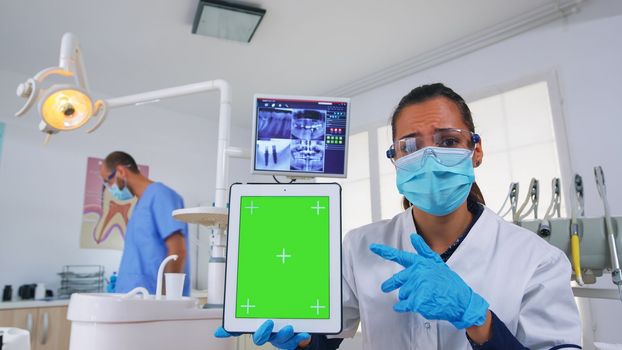 Dentist doctor showing tablet with green screen, explaining dental radiography and diagnosis for teeth infection. Stomatology specialist with face mask pointing at mockup, copy space, chroma display