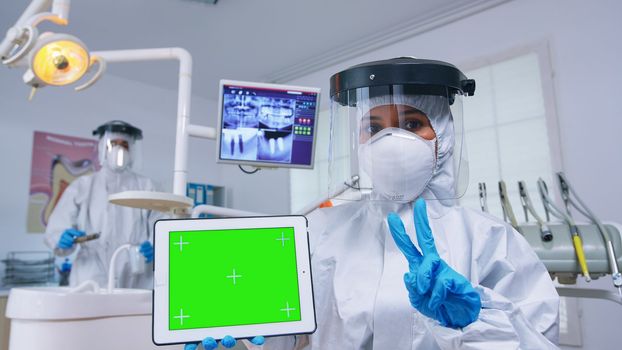 Patient pov of dentist in ppe suit explaining dental radiography and diagnosis for teeth infection using tablet with green screen. Stomatology specialist pointing at mockup, copy space, chroma display