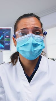 Patient point of view to dentist in protective mask holding tools examining person with toothache sitting on stomatological chair while nurse preparing tools for surgery.