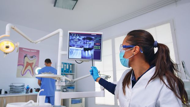 Dentistry doctor talking about dental surgery showing x-ray on monitor unit, patient pov. Stomatology specialist wearing protective face mask pointing at teeth radiography in stomatological clinic