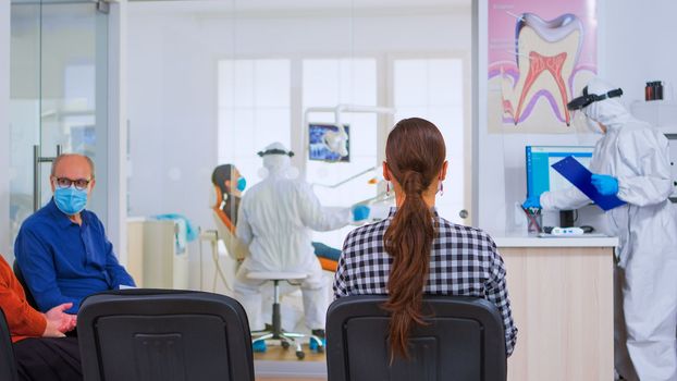 Dental nurse with protection suit holding clipboard leaving reception while patients sitting in waiting room talking keeping distance. Assistant and dentist doctor wearing overall, face shield, gloves