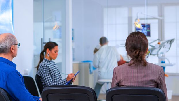 Back view of woman filling in dental form sitting on chiar in waiting room preparing for dental implants while doctor exemination patient in background. Crowded professional orthodontist office.